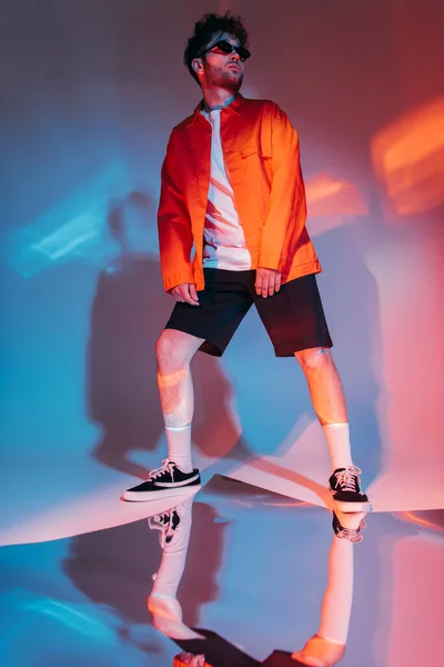 Full length of curly young man in sunglasses posing while looking away on grey with colorful light — Fotografia de Stock