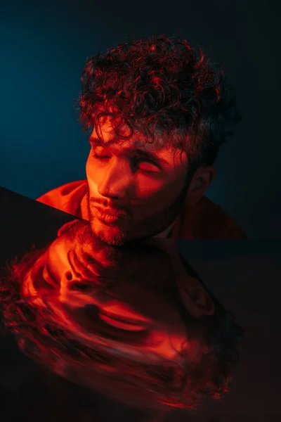 Curly and stylish man in orange shirt posing with closed eyes near mirror on blue — Fotografia de Stock