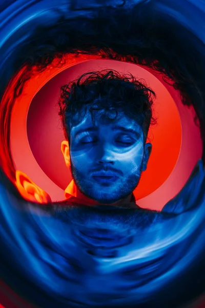 Curly and young man with closed eyed near neon blue circle on red — Photo de stock