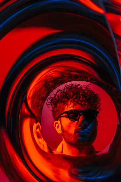 Curly man in sunglasses looking away through futuristic neon circle on red — Fotografia de Stock