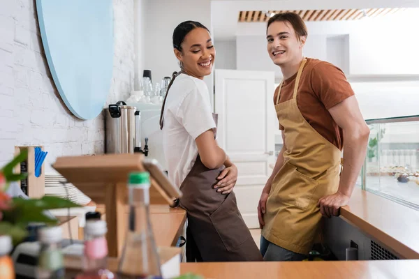 Cheerful multiethnic sellers in aprons standing in sweet shop — Stock Photo