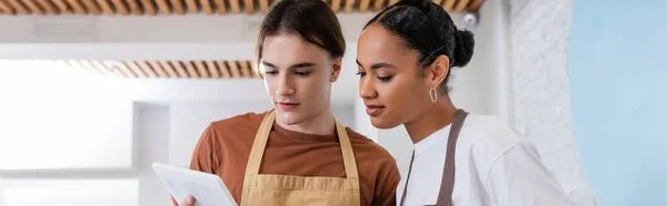 Young interracial sellers in aprons using digital tablet in confectionery, banner — Foto stock