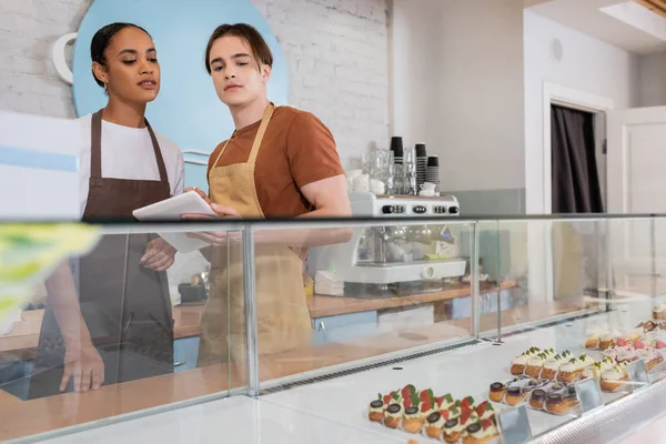 Interracial sellers using digital tablet near eclairs in showcase in sweet shop — Stock Photo