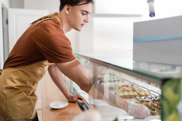 Side view of young seller in glove taking dessert from showcase in sweet shop — Fotografia de Stock