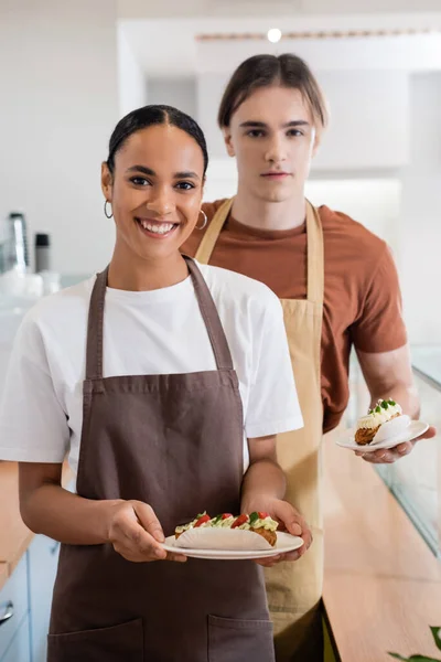 Smiling african american saleswoman holding dessert near colleague in sweet shop - foto de stock