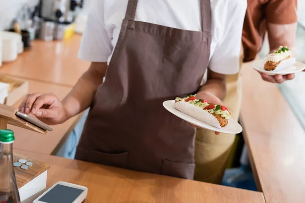 Cropped view of african american seller holding dessert and using digital tablet near colleague in confectionery — стоковое фото