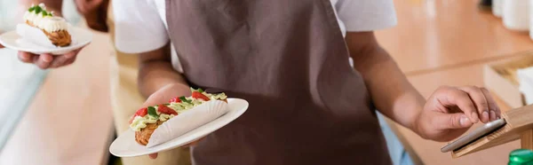 Cropped view of african american saleswoman holding dessert and using digital tablet in sweet shop, banner — Photo de stock