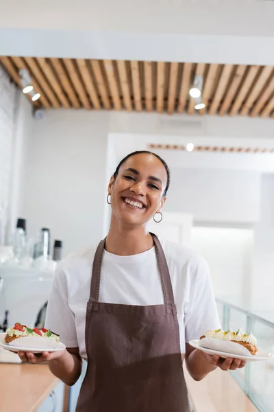 Cheerful african american seller holding eclairs and looking at camera in confectionery — Stockfoto
