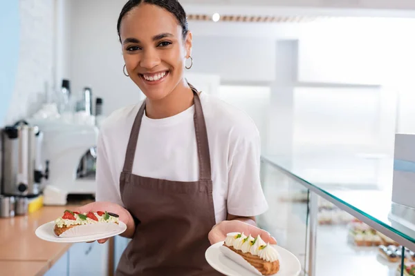 Cheerful african american seller in apron holding eclairs in sweet shop — Stock Photo
