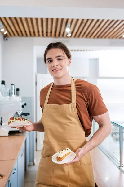 Smiling salesman in apron holding eclairs and looking at camera in sweet shop — Stock Photo