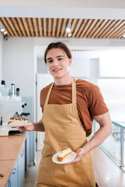 Smiling salesman in apron holding eclairs and looking at camera in sweet shop — стоковое фото