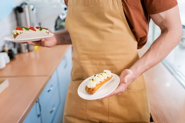 Cropped view of seller in apron holding tasty eclairs in confectionery — стоковое фото