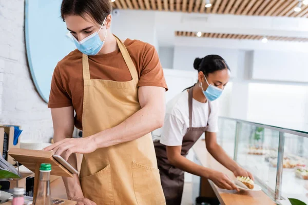 Seller in medical mask using digital tablet near african american colleague with dessert in sweet shop — Stockfoto