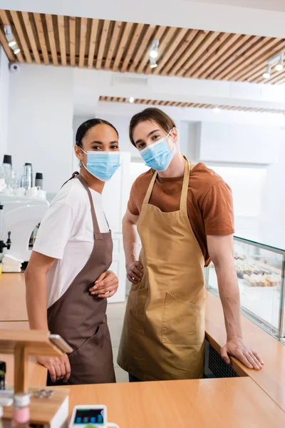 Multiethnic sellers in medical masks looking at camera in sweet shop — Photo de stock