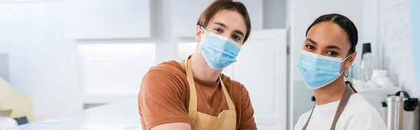 Multiethnische Verkäufer in medizinischen Masken blicken in die Kamera in der Konditorei, Banner — Stockfoto