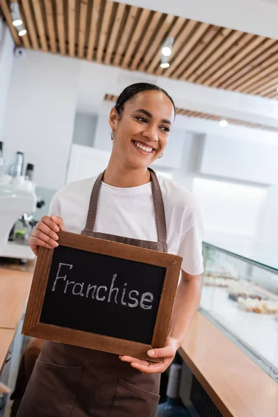 Positive african american seller in apron holding chalkboard with franchise lettering in confectionery — Stock Photo