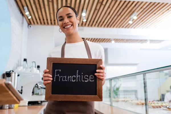 Positive african american seller holding chalkboard with franchise lettering near showcase in sweet shop — Stock Photo