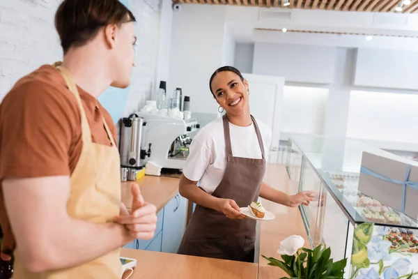 Smiling african american seller holding dessert near showcase and blurred colleague in sweet shop — Foto stock