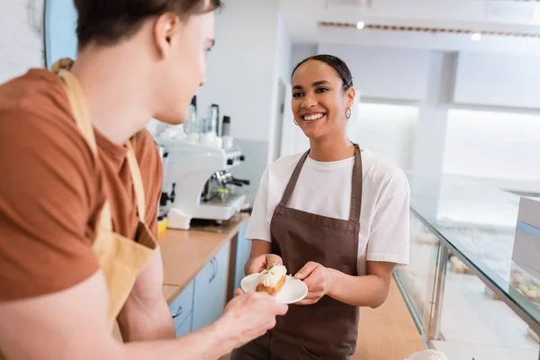 Cheerful african american saleswoman giving elcair to blurred colleague in sweet shop — Foto stock