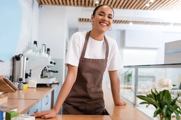 Fröhliche afrikanisch-amerikanische Verkäuferin blickt in die Kamera in der Nähe von Schaufenster und Blume im Süßwarenladen — Stockfoto