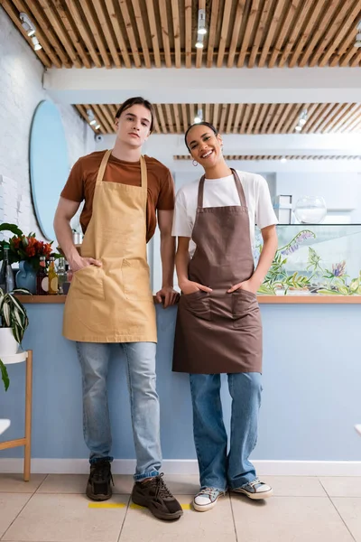 Multiethnic sellers in aprons looking at camera in confectionery — Stock Photo