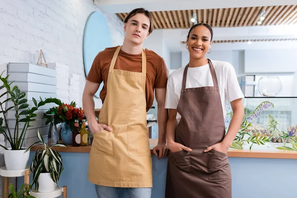 Young interracial sellers in aprons looking at camera near plants in sweet shop — Stock Photo