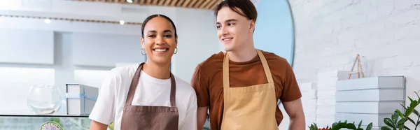 Smiling salesman in apron looking at african american colleague in sweet shop, banner — Stock Photo