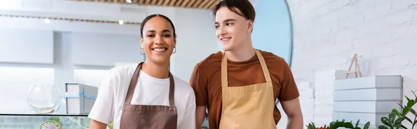 Smiling salesman in apron looking at african american colleague in sweet shop, banner - foto de stock