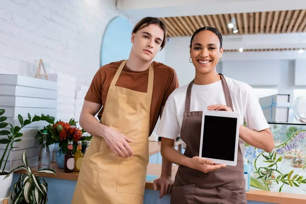 Positive african american seller showing digital tablet near colleague in sweet shop — Stock Photo