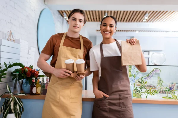 Cheerful multiethnic sellers holding coffee to go and paper bag in confectionery — Foto stock