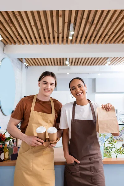 Cheerful multiethnic sellers holding coffee to go and paper bag in confectionery - foto de stock