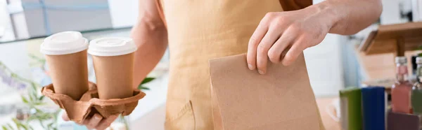 Cropped view of salesman holding paper bag and coffee to go in confectionery, banner - foto de stock