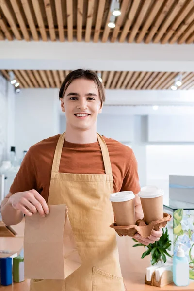 Cheerful young salesman holding coffee to go and paper bag in sweet shop — Stock Photo