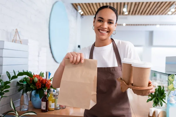 Positiver afrikanisch-amerikanischer Verkäufer mit Coffee to go und Papiertüte im Süßwarenladen — Stockfoto