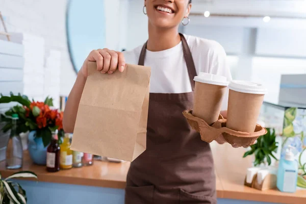 Cropped view of african american seller holding paper bag and takeaway coffee in confectionery — стоковое фото