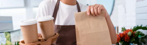 Cropped view of african american worker holding coffee to go and paper bag in sweet shop, banner — Foto stock