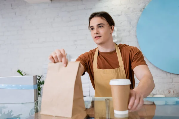 Seller in apron holding paper bag and takeaway drink in confectionery — Fotografia de Stock