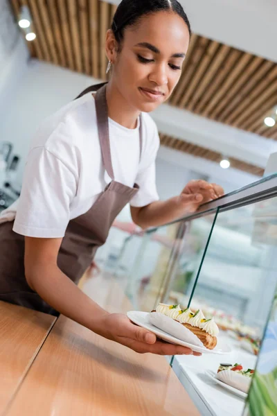 African american saleswoman in apron holding eclair near showcase in confectionery — Photo de stock