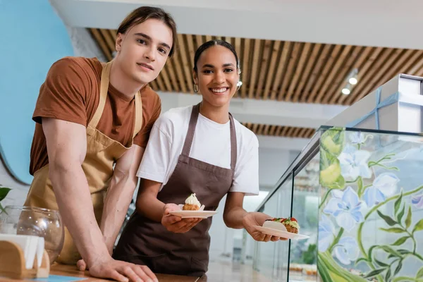 Cheerful interracial sellers holding eclairs and looking at camera in confectionery — Photo de stock