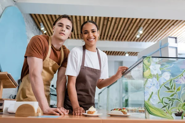 Positive multiethnic sellers in aprons looking at camera near eclairs in sweet shop — Foto stock