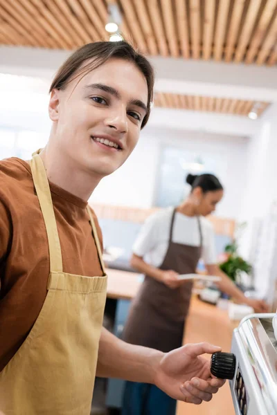 Smiling barista in apron looking at camera near coffee machine in sweet shop — Stock Photo