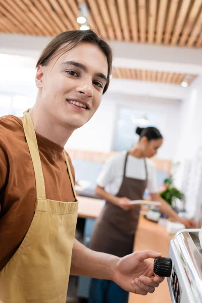 Smiling barista in apron looking at camera near coffee machine in sweet shop — стоковое фото