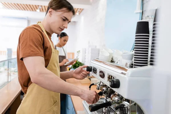 Young barista in apron using coffee machine near blurred african american saleswoman in confectionery — Foto stock
