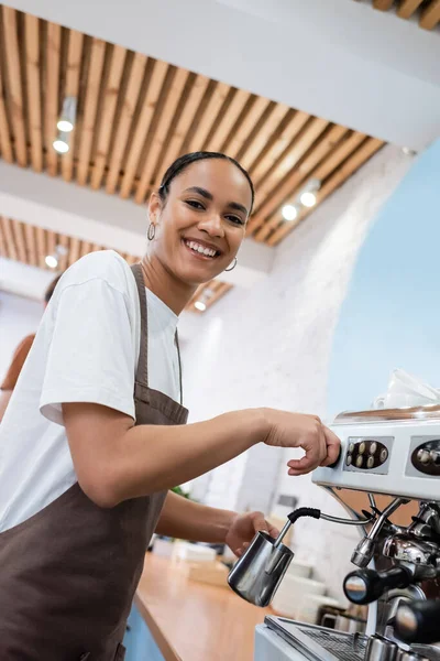Low angle view of smiling african american barista making coffee and looking at camera in confectionery — Stockfoto