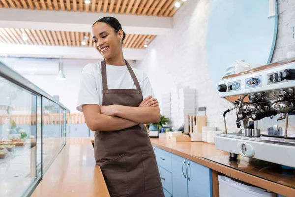 Smiling african american barista looking at showcase near coffee machine in confectionery — стоковое фото