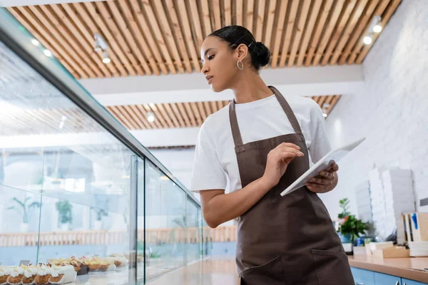 Low angle view of african american saleswoman with digital tablet looking at showcase in sweet shop — Photo de stock