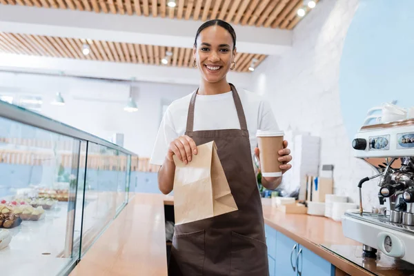 Positive afrikanisch-amerikanische Verkäuferin hält Getränke zum Mitnehmen und Papiertüten in Süßwaren — Stockfoto
