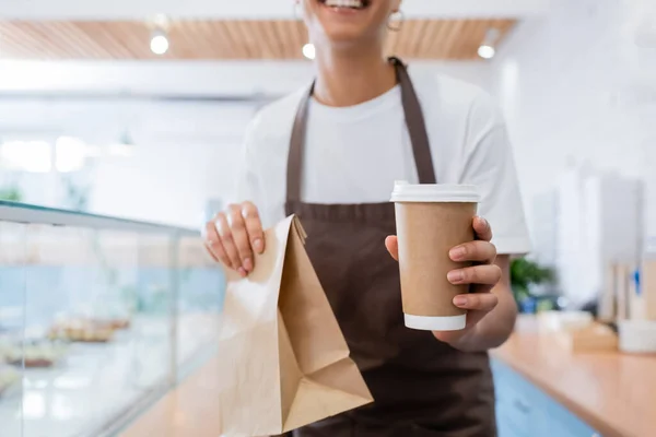 Cropped view of coffee to go and paper bag in hands of blurred african american saleswoman in sweet shop — стоковое фото