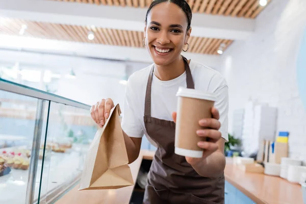 Cheerful african american seller holding paper bag and cup while looking at camera in confectionery — Photo de stock