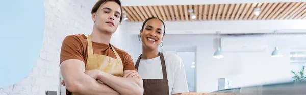 Smiling interracial sellers in aprons looking at camera in confectionery, banner - foto de stock